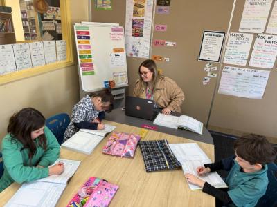 A teacher sits working on a laptop, on either side sit students writing in their workbooks