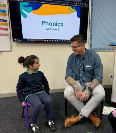 Somerville Rise principal Matt Jackson sits next to a student in the classroom, behind them a whiteboard reads phonics session 2