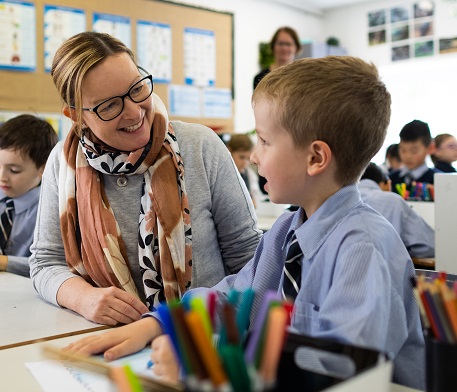 A teacher and a primary school boy face each other at a desk in a classroom. The boy has his work in front of him. The teacher is smiling. 