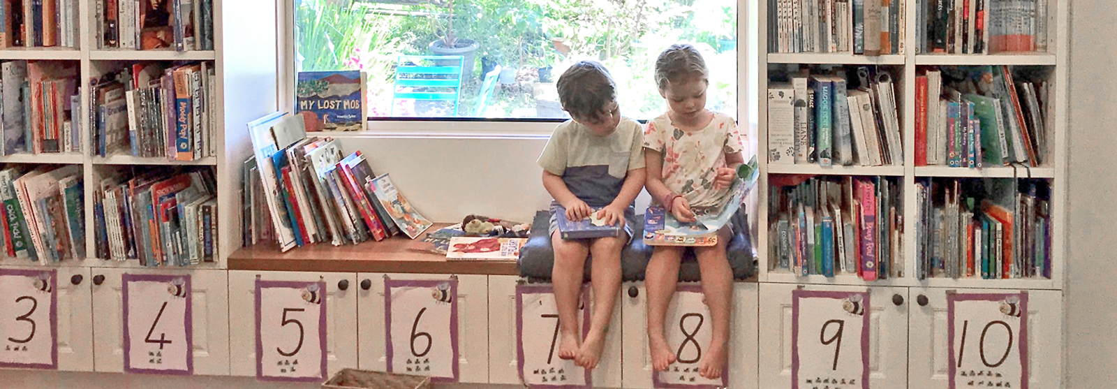 A boy and a girl sit on a window seat looking at a book. Shelves of books surround them on the left and right.