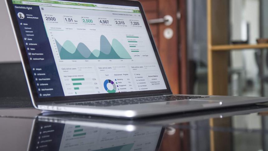 A laptop sits open on a mirror-topped desk. On the screen are a number of charts and graphs. The words and numbers are not legible but look like a school improvement dashboard.