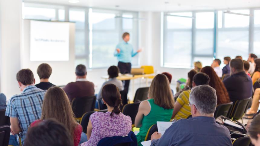 A lecturer stands in front of a room full of people sitting and watching them