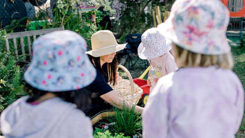 Two female children, wearing hats, watch an educator and another child working in a vegetable garden at Canning Vale Prekindy. 