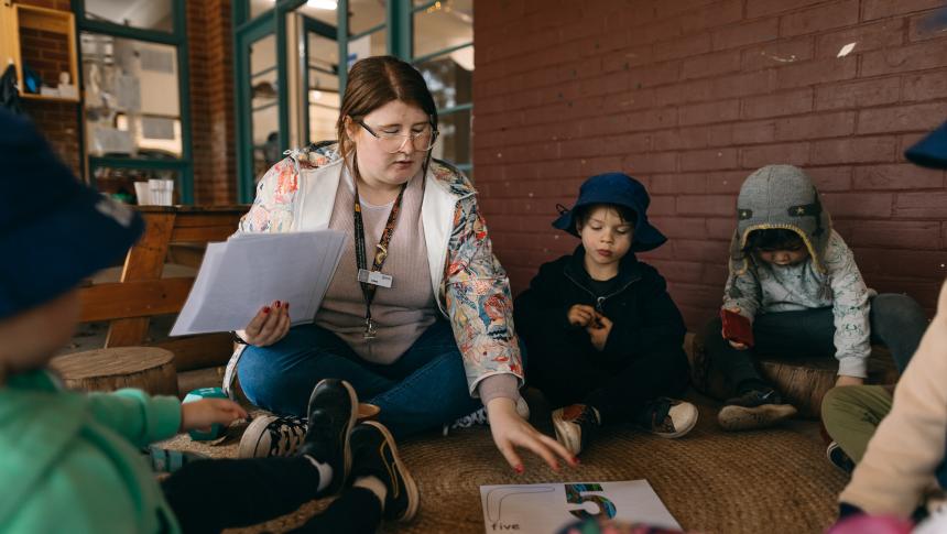 teacher sitting on the ground with young children looking at a number five