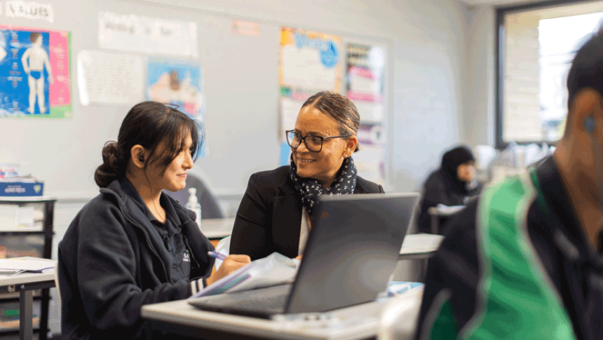 A student works at a desk while a teacher crouches beside her