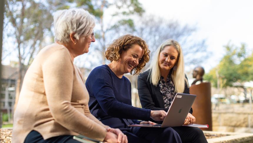 Three professional women sitting outdoors and looking at a laptop.