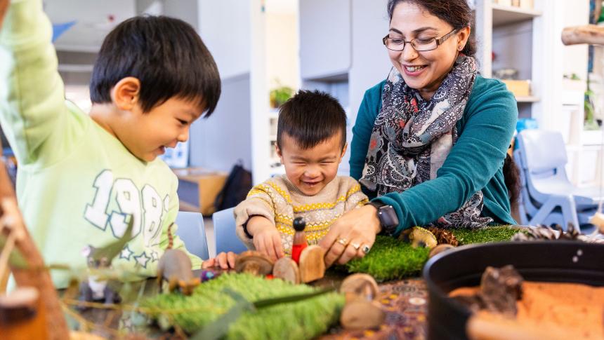 Teacher or educator playing with wooden figures with 2 children at table.