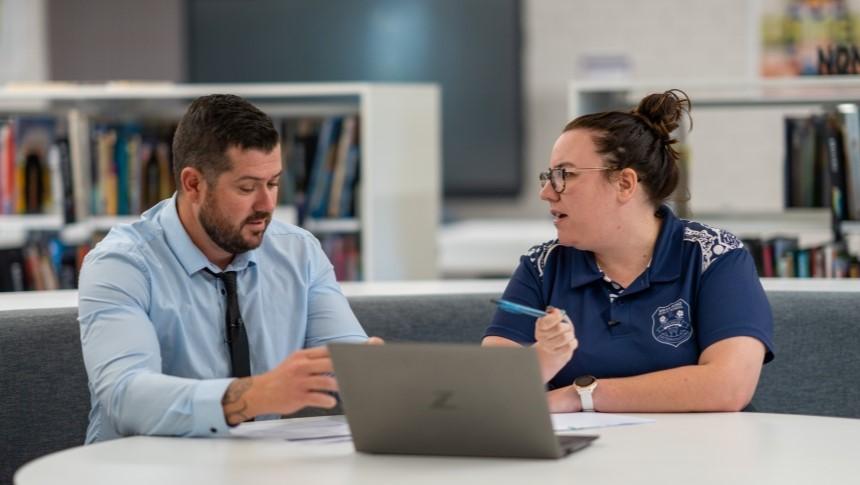 Two teachers sitting at a table in a library having a discussion, with a laptop open in front of them