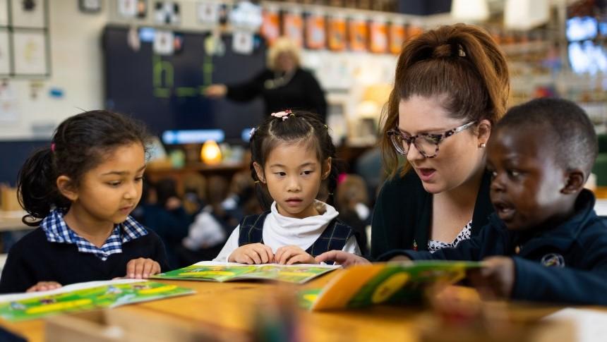 A teacher sitting at a low desk with several primary school students helping them with reading