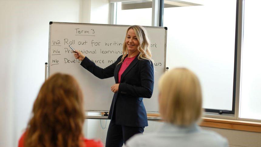 An educator giving a presentation to two other educators, standing at a whiteboard with plans for several weeks of Term 3