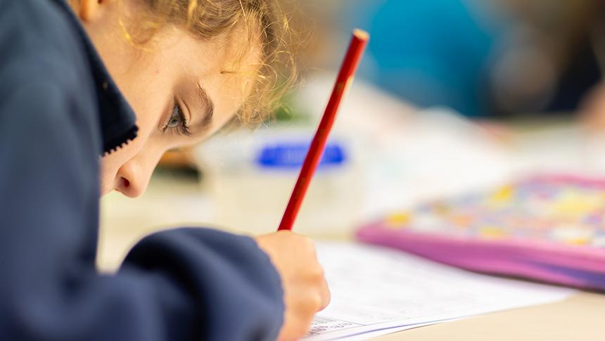Primary school student bent over desk writing with on paper pencil.