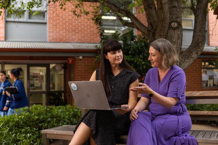 Two female teachers have a conversation while viewing a laptop in the school yard.
