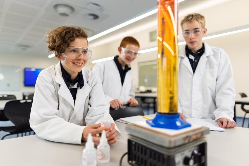 3 male students happily conducting a science experiment wearing lab cots and goggles.