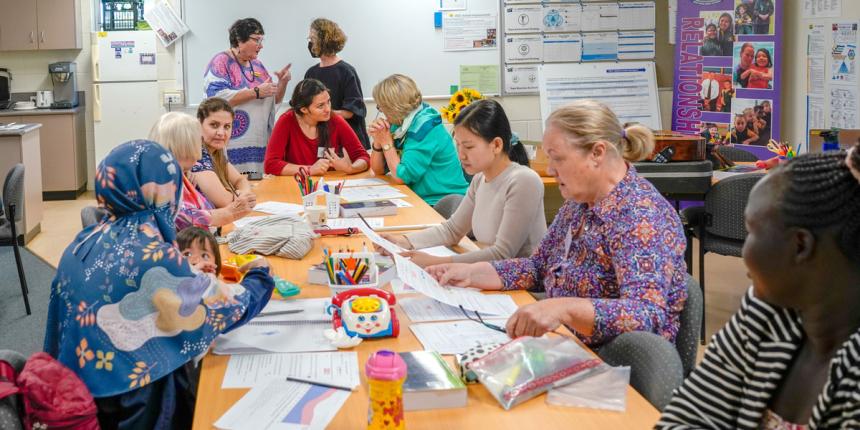 A group of eight woman from different ethnic backgrounds sit around a table talking to one another. Another two women stand in the background, having a conversation.