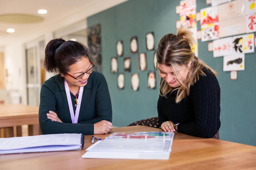 High school student sitting at desk with teacher discussing work.