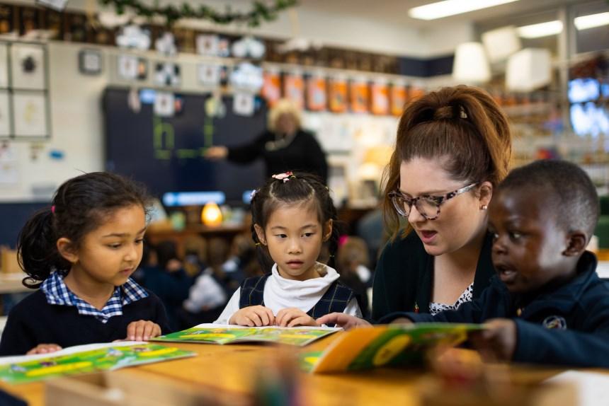 A teacher sitting at a low desk with several primary school students helping them with reading